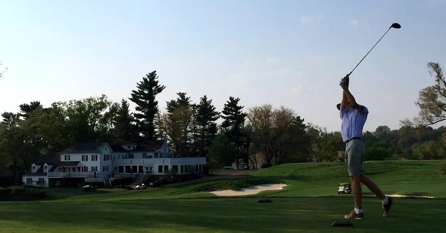 A golfer finishing a drive in front of a clubhouse at sunset